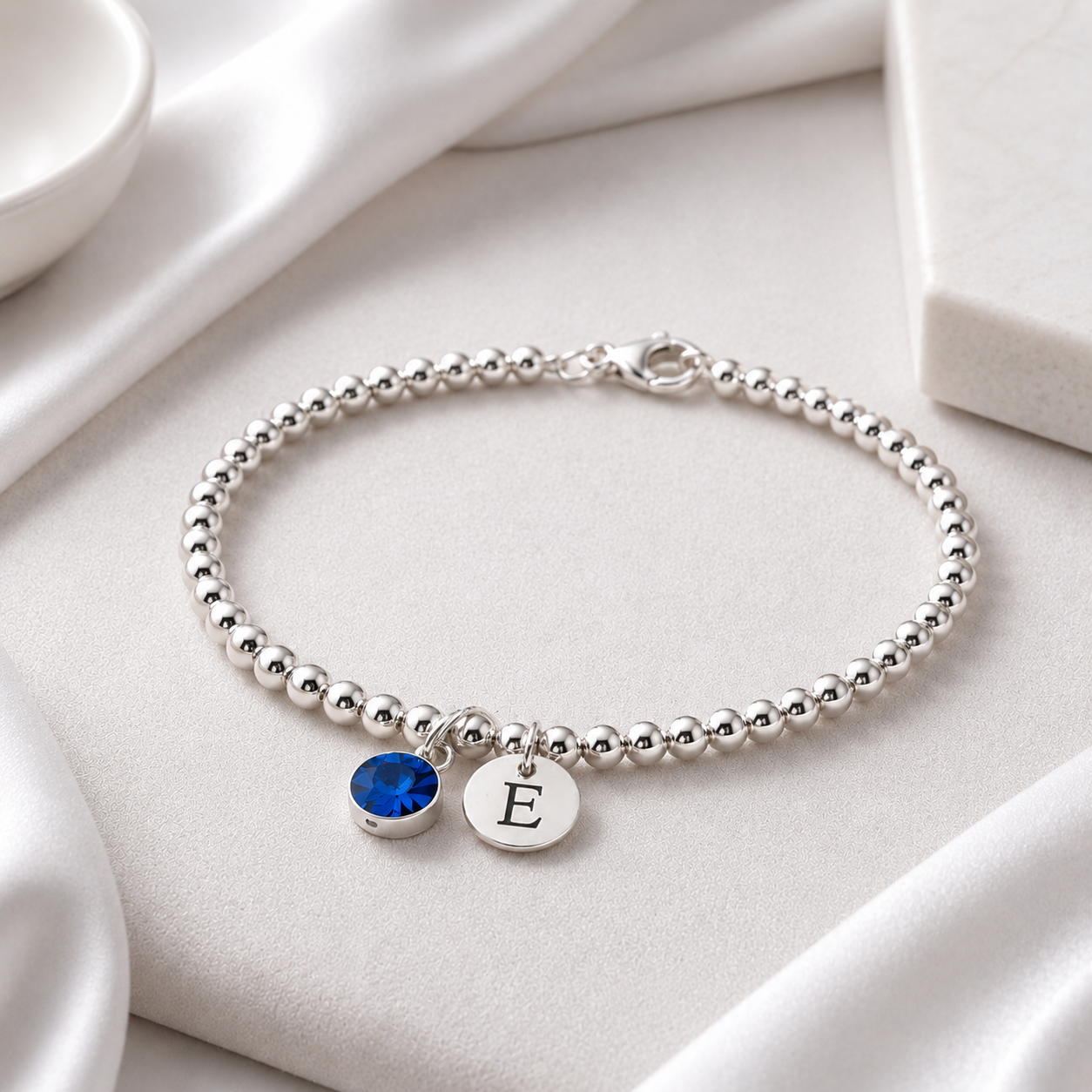  and a silver disc charm engraved with the letter 'E', displayed on a white satin fabric background.